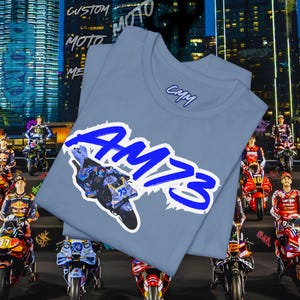 May include: Two light blue t-shirts with a crew neck are displayed. The shirts feature a blue and white graphic of a motorcycle and the text "AM73". The shirts also have the text "CMM" and "CUSTOM MOTO" in white.