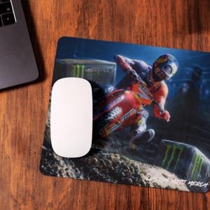 May include: A custom motocross mousepad featuring a red, white, and blue motocross rider on a dirt bike. The rider is wearing a helmet and goggles. The mousepad has a black background with a Monster Energy logo in the bottom right corner. The text "Custom Moto Merch" is printed in white at the bottom of the mousepad.