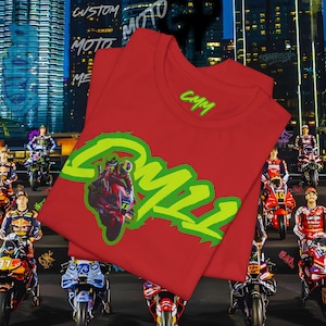 Puede incluir: Camiseta roja con un diseño de motocicleta y el texto "CMM" en verde. La camiseta está doblada. El fondo muestra un paisaje urbano y corredores de motos.