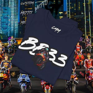 May include: Navy blue t-shirt with a graphic of a motorcycle racer and the text "BR33" in white. The shirt also features the initials "CMM" in white. The background shows a cityscape and several motorcycles.