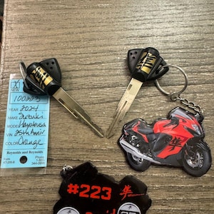 May include: Two black keys with gold lettering and a black motorcycle keychain with red and black detailing. The keychain has the text "#223" and "25th Anniversary" on it.