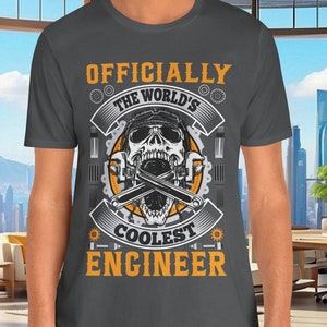 以下が含まれることがあります： 暗いグレーのTシャツに、ヘルメットをかぶってレンチを持ったドクロのグラフィックデザインが施されています。Tシャツには「Officially The World's Coolest Engineer」という文字が書かれています。