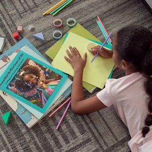 May include: A young person is coloring on a sheet of yellow paper with a purple pencil. The person is lying on a gray carpet with a book titled "Beautiful Reflections of Me: A Magical Coloring Adventure" on the floor next to them.