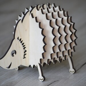 May include: A wooden hedgehog figurine with a layered design. The figurine is made of light-colored wood with dark brown edges, featuring a smiling face and small legs. The hedgehog's body has a spiky appearance.