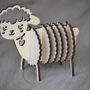 May include: A wooden sheep figurine with a layered design. The sheep has a light-colored body and a darker outline, with a heart-shaped cutout. The figurine is standing on four small legs.