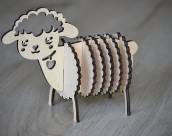 May include: A wooden sheep figurine with a layered design. The sheep has a light-coloured body and a darker outline, with a heart-shaped cutout. The figurine is standing on four small legs.