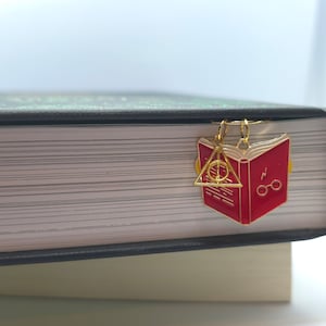 Könnte beinhalten: Ein goldener Buch-Anhänger mit einem roten Emaille-Buch und einem goldenen Blitz- und Brillen-Design. Der Anhänger ist an einem Buch mit schwarzem Einband befestigt.