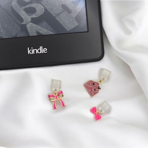 Könnte beinhalten: Drei rosa und goldene Charms vor weißem Hintergrund. Die Charms sind eine Schleife, ein herzförmiger Umschlag und eine weitere Schleife. Die Charms sind an durchsichtigen Plastikclips befestigt.