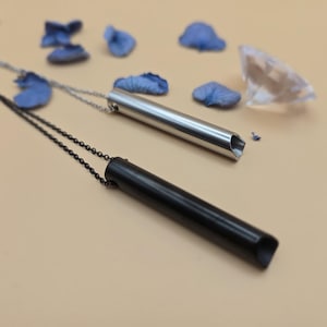 Könnte beinhalten: Zwei Halsketten mit Anhänger. Eine Halskette hat einen silbernen Anhänger in Form eines Rohrs mit einem Ausschnitt an einem Ende. Die andere Halskette hat einen schwarzen Anhänger in Form eines Rohrs mit einem Ausschnitt an einem Ende.