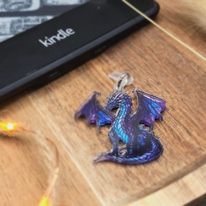 Könnte beinhalten: Ein lila und blauer Drachen-Schlüsselanhänger mit ausgebreiteten Flügeln. Der Drache ist aus klarem Acryl mit einem bedruckten Design.