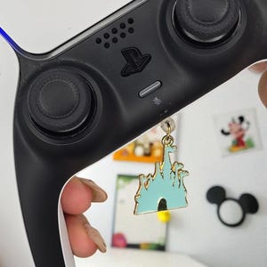 Könnte beinhalten: Ein goldfarbener Anhänger mit einem hellblauen Schlossdesign mit schwarzen Akzenten, der an einem schwarz-weißen Gamecontroller hängt. Der Anhänger ist ein dekoratives Accessoire.