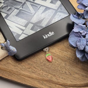 Könnte beinhalten: Schwarzer Kindle E-Reader auf einem Holztablett. Ein kleiner Erdbeer-Anhänger mit roter Emaille und grünem Oberteil hängt an dem Gerät. Getrocknete blaue Blumen sind um den E-Reader angeordnet.