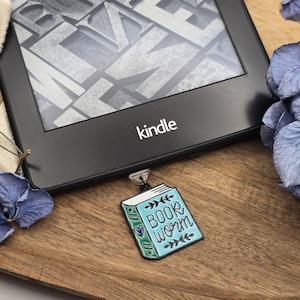Könnte beinhalten: Eine blaue und grüne Emaille-Anstecknadel in Form eines Buches mit den Worten "Book Worm" auf dem Cover. Die Anstecknadel ist an einem schwarzen Kindle E-Reader befestigt.