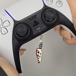 Könnte beinhalten: Ein weiß-schwarzer Videospiel-Controller mit einem silbernen Anhänger. Der Anhänger ist ein Messer mit Totenkopf-Design, mit roter und weißer Emaille. Der Controller hat schwarze Joysticks und Tasten.