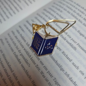 Może przedstawiać: Złoty zakładka z niebiesko-białym emaliowanym brelokiem w kształcie książki. Brelok ma okulary i cyfrę "5" na okładce. Zakładka jest otwarta i ma przymocowany złoty pierścień.
