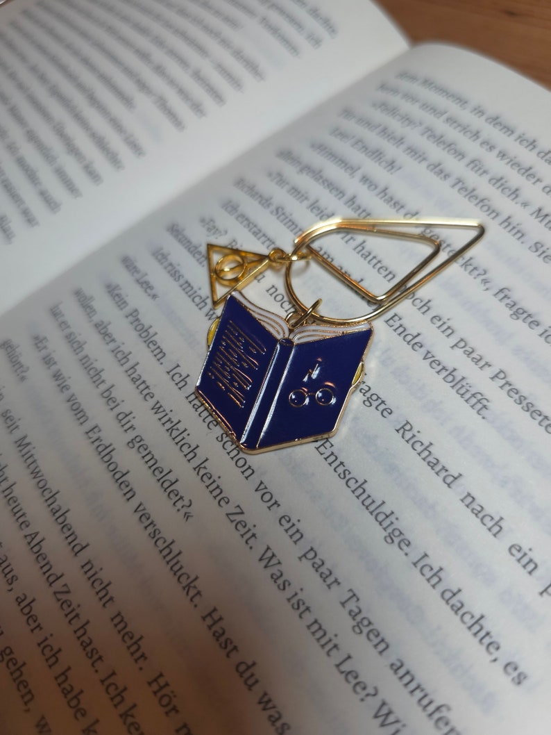 Może przedstawiać: Złoty zakładka z niebiesko-białym emaliowanym brelokiem w kształcie książki. Brelok ma okulary i cyfrę "5" na okładce. Zakładka jest otwarta i ma przymocowany złoty pierścień.