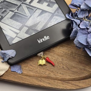 Könnte beinhalten: Ein goldener Buch-Anhänger und ein roter Chili-Anhänger, die an einem schwarzen Kindle E-Reader befestigt sind. Der E-Reader liegt auf einer Holzoberfläche.
