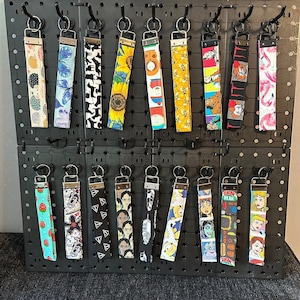 May include: A display of 18 colorful keychains with various designs, including cartoon characters, animals, and pop culture references. The keychains are hanging on a black pegboard.