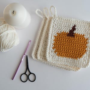 May include: Three crocheted pot holders with a pumpkin design. The pot holders are white with a brown stem and orange pumpkin. The pot holders are made with yarn and a crochet hook.