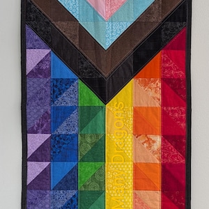 Op de afbeelding: Een gewatteerde wandhanger met een Pride-vlagontwerp. Het bovenste gedeelte heeft een chevronpatroon in roze, lichtblauw, bruin en zwart. Het onderste gedeelte is een regenboog met de tekst "Sexy May Dragons" in geel.