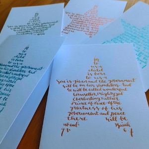 Può includere: Una collezione di biglietti bianchi con calligrafia in vari colori. Ogni biglietto presenta un disegno a forma di stella riempito con il testo di Isaia 9:6. La calligrafia è nelle tonalità del verde acqua, dell'arancione e dell'oro.