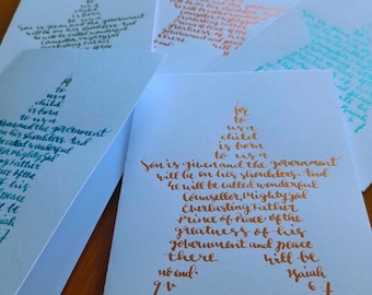 Puede incluir: Un conjunto de tarjetas blancas con caligrafía en varios colores. Cada tarjeta presenta un diseño en forma de estrella lleno de texto de Isaías 9:6. La caligrafía está en tonos de verde azulado, naranja y dorado.