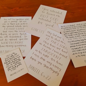 May include: Five white paper prints featuring black hand-lettered calligraphy of Bible verses. The texts include Genesis 50:20, Matthew 10:29-31, Daniel 6:22, Proverbs 3:5-6, and Psalm 23. Each print displays distinct religious quotes written in a decorative script.