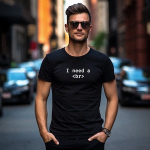 Op de afbeelding: Zwart T-shirt met ronde hals met de tekst "I need a <br>". Het T-shirt wordt gedragen door een persoon die een zonnebril en een horloge draagt. De persoon staat op een stadsstraat.