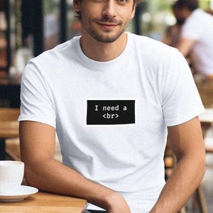 Puede incluir: Camiseta blanca con el texto "I need a <br>" en un rectángulo negro. La camiseta la lleva una persona. Se ve una taza y un platillo blancos sobre una mesa de madera.