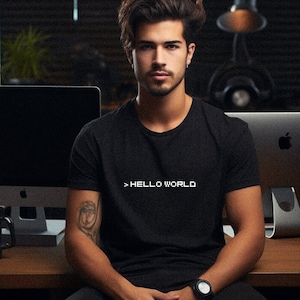 May include: A man wearing a black t-shirt with the text "> HELLO WORLD" in white lettering. He is sitting in front of a computer screen.