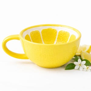 May include: A vibrant yellow ceramic teacup with a handle, showcasing a lemon slice design inside. The cup is displayed with lemon slices, white flowers, and green leaves, set against a white backdrop.