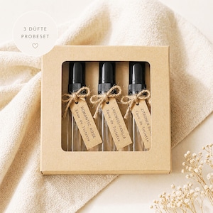 May include: A cardboard box holding three perfume atomisers. Each bottle has a black cap and a tag tied with twine. The tags read "Romantic Rose," "Lavender-Vanilla," and "Zitronen Bou-" with "Eau de Toilette." The box is on a beige cloth.