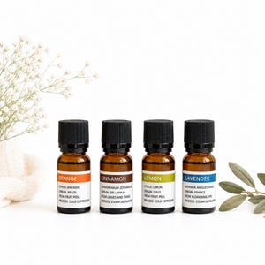 Könnte beinhalten: Vier kleine ätherische Ölflaschen aus braunem Glas mit schwarzen Kappen, beschriftet mit Orange, Zimt, Zitrone und Lavendel. Jedes Etikett enthält den botanischen Namen, die Herkunft und das Extraktionsverfahren. Die Flaschen stehen auf einer weißen Oberfläche.