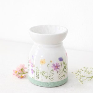 Könnte beinhalten: Weißer Keramik-Diffusor für ätherische Öle mit einem Blumendesign mit lila, gelben und rosa Blumen. Der Sockel hat ein hellgrünes Band. Der Diffusor hat eine breite Öffnung oben.