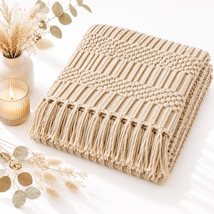 Könnte beinhalten: Eine gefaltete, beigefarbene Strickdecke mit strukturiertem Zickzackmuster und langen Fransen am Rand. Die Decke ist aus dickem Garn gefertigt und zeigt ein sich wiederholendes geometrisches Design in einer weichen, neutralen Farbgebung.