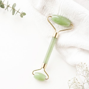 Könnte beinhalten: Eine Gesichtsrolle aus Jade mit goldfarbenen Metallakzenten liegt auf einem weißen Handtuch. Die Rolle ist hellgrün und hat ein glattes, poliertes Aussehen. Eukalyptusblätter und kleine weiße Blüten befinden sich im Hintergrund.