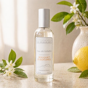Könnte beinhalten: Eine transparente Glasflasche "Eau de toilette" Parfüm mit weißem Deckel. Das Etikett lautet "Bouquet d'Agrumes" und "Fabriqué en France". Die Flasche enthält 100 ml. Eine Zitrone und Blumen sind im Hintergrund.