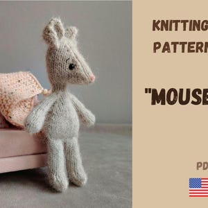 以下が含まれることがあります： ピンクの鼻と黒い目の編み物グレーマウスのおもちゃが、毛布と枕が付いた小さなピンクのソファの隣に立っています。画像には、「KNITTING PATTERN」と「MOUSE」のテキスト、PDF記号、およびアメリカ国旗が含まれています。