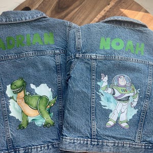 Puede incluir: Dos chaquetas vaqueras con nombres personalizados, "Adrian" y "Noah", y diseños pintados. Una chaqueta presenta un dinosaurio verde, la otra un personaje de dibujos animados de Toy Story.