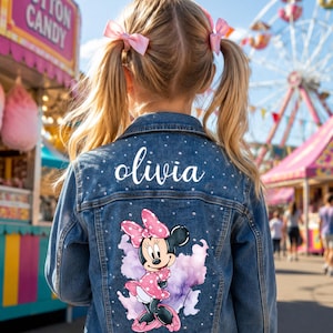 May include: A blue denim jacket featuring a graphic of Minnie Mouse in a pink polka dot dress and bow, surrounded by purple watercolor accents. The name olivia is printed in white script above the graphic, with small rhinestones scattered throughout.