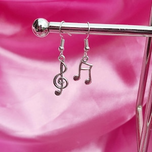 Peut inclure: Boucles d'oreilles argentées avec une clé de sol et une note de musique. Les boucles d'oreilles sont suspendues à un présentoir en métal argenté sur un fond rose. Accessoire pour les mélomanes.