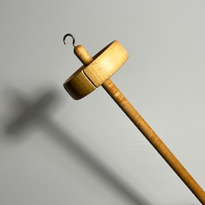Pu&ograve; includere: Un fuso in legno con un volano rotondo e piatto e un lungo e sottile fusto. Il fuso ha un gancio in ottone nella parte superiore. Il legno ha una finitura chiara e naturale. Per la filatura a mano.