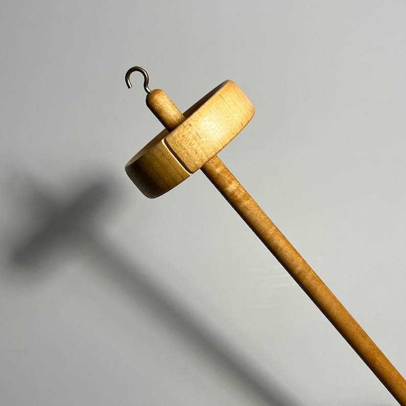 Pu&ograve; includere: Un fuso in legno con un volano rotondo e piatto e un lungo e sottile fusto. Il fuso ha un gancio in ottone nella parte superiore. Il legno ha una finitura chiara e naturale. Per la filatura a mano.