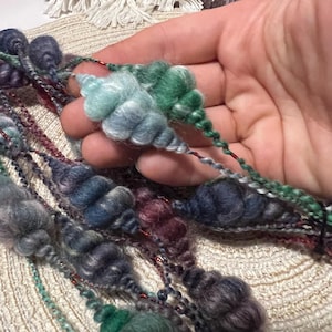 May include: Close-up of colorful hand-spun yarn in shades of blue, green, and purple. The yarn has a textured, bumpy appearance with a metallic thread running through it. The yarn is held in a hand, showcasing its unique texture and color variations.