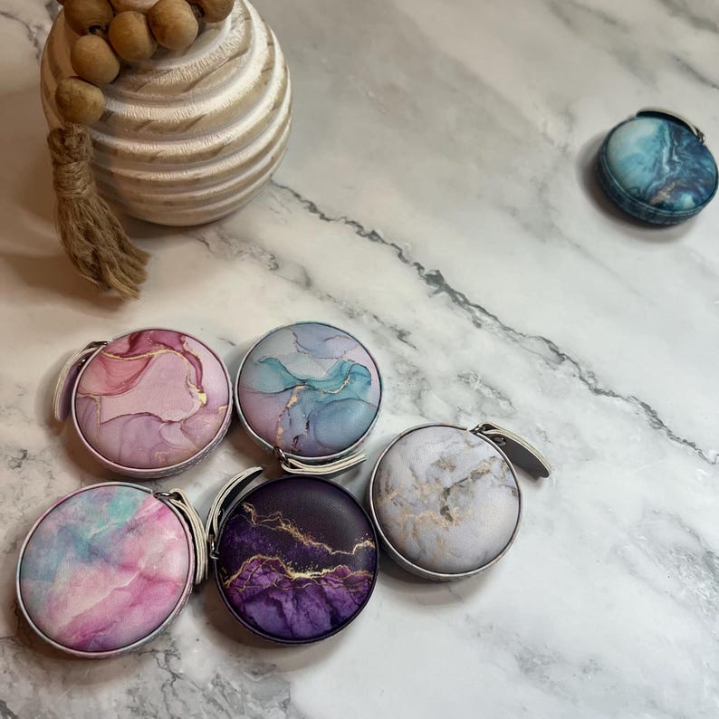 K&ouml;nnte beinhalten: Eine Sammlung runder, dekorativer Ma&szlig;b&auml;nder mit abstrakten Designs in Rosa, Blau, Lila und Wei&szlig;. Jedes Ma&szlig;band hat einen silberfarbenen Griff und ein marmor&auml;hnliches Muster. Der Hintergrund ist eine Marmoroberfl&auml;che.