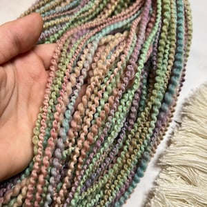 May include: Close-up of a bundle of colorful, textured yarn in pastel shades of pink, green, blue, and beige. The yarn is held in a hand, showcasing its soft, looped texture. A cream-colored fringe is visible in the background.