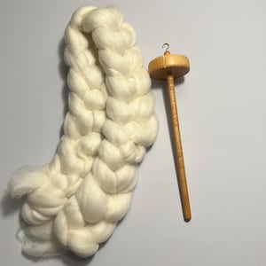 Pu&ograve; includere: Primo piano di una treccia color crema di lana cardata accanto a un fuso in legno. Il fuso ha un fuso rotondo in legno e un lungo e sottile fusto. La lana &egrave; morbida e soffice, ideale per la filatura.