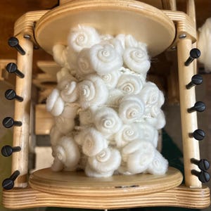 May include: A wooden yarn swift with a large quantity of white, fluffy yarn. The yarn is arranged in a spiral pattern, creating a textured appearance. The swift has a natural wood finish and black hardware.