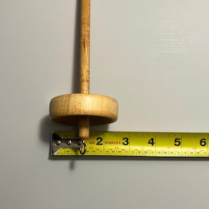 Pu&ograve; includere: Un fuso di legno con un fuso rotondo e un lungo albero. Il fuso &egrave; marrone chiaro e misura circa 5 cm di lunghezza. Un metro a nastro giallo &egrave; posto accanto al fuso.