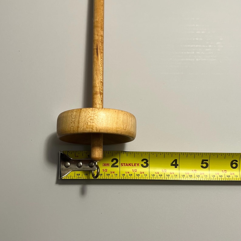 Pu&ograve; includere: Un fuso di legno con un fuso rotondo e un lungo albero. Il fuso &egrave; marrone chiaro e misura circa 5 cm di lunghezza. Un metro a nastro giallo &egrave; posto accanto al fuso.